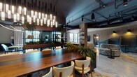 Located at 714 N Racine Ave in Chicago's vibrant West Town neighborhood, this thoughtfully designed single-floor amenity space masterfully blends industrial aesthetics with refined, contemporary interiors. Exposed ceiling infrastructure and dark overhead elements contrast beautifully with warm wood accents, polished concrete floors, and soft textile finishes throughout.

The space is organized into several distinct yet flowing zones. A striking communal dining area anchors one section, featuring a substantial solid-wood table surrounded by cream upholstered chairs and illuminated by a dramatic cascade of pendant lighting. Lush greenery, including large fern arrangements integrated into the furniture, adds a vibrant, organic energy to the space.

Adjacent to the dining area, a full-service kitchen bar with deep blue cabinetry and open display shelving offers a polished hospitality-style setup complete with screens for entertainment. A lounge zone transitions seamlessly nearby, offering generous light-gray sofas, a slatted wood ceiling detail, and a curved architectural partition that adds visual interest.

Tucked-away booth seating with arched alcoves and warm pendant lighting provides intimate gathering options, while private workspaces with floor-to-ceiling windows offer views of an outdoor terrace. The overall ambiance is sophisticated yet welcoming, balancing community-focused gathering with quieter, more focused areas.
