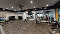 Located at 1443 Devon Ln in Harrisonburg, Virginia, this single-floor fitness facility offers a well-appointed and thoughtfully designed space for a full range of exercise activities. The main strength training area features a generous open floor plan with textured carpet tile flooring, recessed lighting, and a dark-painted ceiling that lends a modern, energetic atmosphere. This zone is outfitted with a robust selection of cable machines, selectorized strength equipment, free weights including a full dumbbell rack, weight benches, and a prominent digital interval timer — all arranged to maximize usability and traffic flow. Large floor-to-ceiling mirrors line multiple walls, enhancing the sense of space and supporting proper form during workouts. A striking multicolored accent wall adds a bold visual element to the strength area, while natural light filters in through windows along one wall. Adjacent to the main floor is a dedicated functional training studio featuring light hardwood-style flooring, wall-mounted flat-screen TVs, full-length mirrors, TRX suspension trainers, and plyo boxes — ideal for group or individual training sessions. The facility also includes a clean hallway corridor with a water fountain station, a unisex restroom, and storage shelving, ensuring members have convenient access to essential amenities throughout their visit.