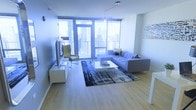 This contemporary single-floor urban residence is defined by its striking floor-to-ceiling windows that frame sweeping city skyline views, flooding the interior with abundant natural light throughout the day. The open-plan living area serves as the centerpiece of the space, showcasing clean-lined modern design with a neutral, sophisticated palette. Light wood flooring extends across the living area, lending warmth and continuity to the space. A large L-shaped gray sectional sofa anchors the room, complemented by a sleek white coffee table and a patterned area rug that adds texture and depth to the layout. A flat-screen television is mounted on a low-profile media console, while a yellow accent chair provides a pop of color and a cozy reading nook near the windows. The expansive glazed doors provide access to a balcony, further enhancing the connection between the interior and the dynamic cityscape beyond. A contemporary pendant light fixture descends from the ceiling, adding an elegant focal point above the living area. The overall ambiance is bright, airy, and sophisticated, reflecting a modern urban lifestyle with an emphasis on open space, natural light, and panoramic city views.
