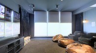 This single-floor space is purpose-built for entertainment and media viewing, designed with a dark, cinematic aesthetic throughout. The room is finished in deep charcoal and navy tones across the walls, ceiling, and carpet, creating an immersive, light-controlled environment ideal for screenings or gaming. Three large roller blinds span a wide window wall, offering full light control when drawn, while allowing natural light to filter softly into the space. Directional track lighting mounted to the ceiling provides focused illumination without disrupting the room's moody ambiance. Seating is casual and inviting, with a collection of oversized leather bean bags arranged toward the front of the room, complemented by additional seating along the side. Dark velvet-style curtains flank the window wall, adding a luxurious, theatrical touch. A low wooden bench sits along one wall, providing a practical surface for equipment or accessories. The overall atmosphere is relaxed yet purposefully designed, blending comfort and function into a cohesive entertainment-focused environment that feels both intimate and well-appointed.