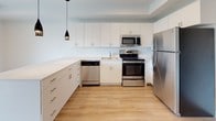 This single-floor unit presents a clean, modern aesthetic carried throughout by light hardwood flooring and a crisp white color palette. The open living area is a standout feature, anchored by large sliding glass doors dressed with vertical blinds that allow natural light to filter in while offering views of the surrounding cityscape and treetops beyond.

The kitchen is thoughtfully designed with an open layout, featuring a generous peninsula countertop with bright white quartz surfaces that double as a breakfast bar. White flat-front cabinetry extends to the upper walls, providing ample storage, while a full suite of stainless steel appliances — including a refrigerator, range, microwave, and dishwasher — adds a polished, functional finish. Sleek black pendant lights hang above the peninsula, offering a subtle contrast to the bright surroundings.

The bathroom maintains the unit's clean design language, with a full soaking tub and shower combination, a white vanity with an integrated sink, and consistent hardwood-style flooring. The overall layout, visible in the 3D overview, flows efficiently, connecting the living, kitchen, bathroom, and utility areas in a compact yet well-organized single-level arrangement.