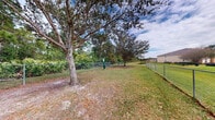 Located at 1900 Bella Vista Way in Port St. Lucie, Florida, this outdoor dog park offers a dedicated, enclosed space for pets to roam and play freely. The area is fully enclosed with chain-link fencing, providing a secure environment for dogs of all sizes. The ground is a mix of grass and natural sandy soil, giving the space a casual, open-air feel typical of Florida's landscape. Several mature oak and pine trees are situated within and around the perimeter, offering natural shade throughout the area — a welcome feature in Florida's warm climate. A waste disposal station is conveniently positioned within the enclosure, promoting cleanliness and responsible pet ownership. The park enjoys open sky views with partial cloud cover visible above, contributing to a bright and airy outdoor atmosphere. Surrounding the fenced area, lush greenery and additional trees provide a natural buffer, while views of the neighboring residential community are visible in the background. The overall setting is practical and functional, designed with the comfort and safety of pets and their owners in mind, making it a valuable community amenity within the Bella Vista Way neighborhood of Port St. Lucie.