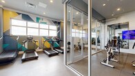 This fitness center offers a comprehensive workout facility designed to accommodate a wide range of exercise needs. The space is organized into distinct functional zones, creating an efficient and energizing environment for users of all fitness levels. A glass-enclosed cycling studio sits at the heart of the layout, providing a defined space for indoor cycling classes while maintaining visual openness through floor-to-ceiling glass partitions and a glass door entry. The cardio zone is highlighted by a bold geometric mural in teal, yellow, and gray tones that spans the accent walls, complemented by large windows that allow abundant natural light to fill the space. This area features treadmills, elliptical machines, and a rowing machine. The strength training zone is well-appointed with a cable machine, a power rack, battle ropes, kettlebells, and an extensive medicine ball rack. Full-length mirrors and a mounted flat-screen television enhance the functionality of this area. Throughout the facility, durable low-pile flooring and recessed lighting contribute to a clean, modern aesthetic. The overall design balances vibrant visual energy with practical, organized workout spaces, making it a fully functional and visually appealing fitness destination.