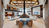 Located at 15621 Bovis Way in Fort Worth, Texas, this impressive clubhouse delivers a grand, resort-caliber experience across a generously sized single floor. The architectural design centers on a dramatic double-height atrium anchored by a stunning suspended circular fireplace, encircled by woven ottomans and plush lounge seating. Above, an elaborate coffered ceiling with warm wood beams draws the eye upward, reinforcing the sense of volume and craftsmanship throughout the space.

The open-plan layout flows seamlessly between distinct social zones. A lounge area features a full-height marble accent wall with an integrated flat-screen display, flanked by rich wood paneling and curated artwork. A billiards table and additional seating vignettes offer recreational gathering spots, while a sleek bar and kitchen area showcases a large marble waterfall island beneath elegant pendant lighting, complemented by floor-to-ceiling windows that invite natural light and outdoor views.

An upper-level mezzanine overlooks the main floor via modern metal railings, providing additional gathering and workspace areas with warm wood-clad walls. Large-format porcelain tile flooring runs throughout, unifying the various zones. The overall ambiance balances contemporary sophistication with warm, textural materials, making this clubhouse a distinctive and welcoming community hub.