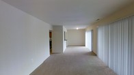 This single-floor space features an open layout with a spacious living area finished in neutral, cream-toned walls and wall-to-wall carpet in a warm beige hue. The living area is generously proportioned, offering a clean, open feel that lends itself well to a variety of furniture arrangements. A flush-mount ceiling light provides overhead illumination, while a large window dressed with vertical blinds along the right wall allows natural light to filter into the space, brightening the interior throughout the day.

A partial wall divides the living area from an adjacent zone, which leads toward the kitchen. The kitchen is visible in the background, featuring dark wood cabinetry and what appears to be a countertop pass-through or opening that maintains a visual connection between the two areas. The transition between the carpeted living space and the kitchen area suggests a shift to hard flooring in that zone.

The overall ambiance is clean, minimalist, and functional, with simple architectural lines and an absence of decorative detailing that makes the space feel open and adaptable. The neutral palette throughout creates a cohesive, understated environment well-suited for customization.