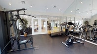 This single-floor home gym is thoughtfully designed to deliver a comprehensive fitness experience in a bright, well-appointed setting. The space features vaulted ceilings that contribute to an open, airy atmosphere, while recessed track lighting ensures consistent, even illumination throughout. A full wall of floor-to-ceiling mirrors visually expands the room and allows for proper form monitoring during workouts. The rubber-floored exercise area is equipped with a multi-function cable machine, a treadmill, and a stationary bike, providing options for both strength training and cardiovascular exercise. A rowing machine rounds out the equipment selection, making this a versatile training environment. The space is anchored by elegant glass-paned French doors that open directly into the adjacent living area, maintaining a visual connection to the rest of the home and allowing natural light to flow in. Decorative potted plants are placed throughout, softening the utilitarian character of the gym and adding a natural, welcoming element. The clean white walls and dark flooring create a crisp, professional aesthetic that is both functional and visually appealing.
