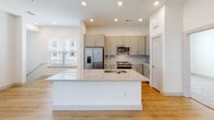 This single-story space in Round Rock, Texas is designed with a clean, contemporary aesthetic throughout. The open-concept layout connects the main living area to a well-appointed kitchen, which serves as a central hub of the home. The kitchen features a large island with a granite countertop and undermount double sink, gray shaker-style cabinetry, a herringbone tile backsplash, stainless steel appliances including a French door refrigerator, and recessed lighting that keeps the space bright and functional. Light wood-tone flooring runs throughout the main living and kitchen areas, contributing to an airy, cohesive feel.

A hallway leading from the main living area includes built-in open shelving, offering practical storage and display space. The bedrooms are carpeted for comfort and include ceiling fans. Each bedroom connects to a bathroom finished with granite-topped vanities, gray cabinetry, and wood-tone flooring. The primary bathroom features a glass-enclosed walk-in shower with tile surround. Natural light enters through multiple windows fitted with blinds. The main living area also opens to a private balcony through sliding glass doors, extending the usable space outdoors.
