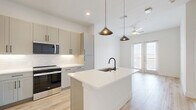 Located at 541 J Pl in Plano, TX, this thoughtfully designed single-floor residence offers a clean, modern aesthetic throughout its well-appointed layout. The kitchen serves as a functional centerpiece, featuring light gray shaker cabinets with matte black hardware, white quartz countertops, a large island with an undermount sink and black gooseneck faucet, stainless steel appliances including a French door refrigerator, range, and microwave, and a crisp white subway tile backsplash. Warm light wood-style flooring and stylish pendant lighting complete the space. A frosted glass sliding door leads to an adjacent area, allowing natural light to flow freely. The bedroom is generously sized with soft wall-to-wall carpeting and a striking bank of floor-to-ceiling windows with transom panels above, flooding the room with natural light and offering neighborhood views. The room connects directly to a dedicated closet and the bathroom. The bathroom is elegantly finished with a double-sink vanity, gray cabinetry, matte black fixtures, large mirrors with modern lighting, and marble-look tile surrounding the walk-in shower. Light wood flooring carries through, tying the space together with a cohesive, contemporary feel.