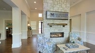 Mount Victor Olde Towne is a thoughtfully designed single-floor community space located in Olde Towne, US. The interior immediately captures attention with a grand floor-to-ceiling stone fireplace serving as the central architectural focal point. Crafted from light-toned natural stone, the fireplace is accented by a rustic reclaimed wood mantel, creating a compelling contrast between refined and organic materials. A sleek linear gas insert adds a modern touch to this classic design element, while the prominent Mount Victor Olde Towne signage mounted above lends the space a polished, branded identity. Rich hardwood flooring runs throughout, grounding the space with warmth and continuity. Soaring ceilings amplify the sense of openness, and strategically placed windows invite abundant natural light, brightening the neutral-toned walls. The seating area features patterned upholstered armchairs in soft blue hues, complemented by light-colored accent tables that reinforce a coastal-inspired aesthetic. A secondary area visible in the background includes additional furnishings and décor, suggesting a multifunctional layout suited for both welcoming guests and facilitating community gatherings. The overall ambiance is warm, sophisticated, and approachable.