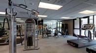 This modern fitness center is designed to support a comprehensive workout experience within a thoughtfully laid-out single-floor space. The facility is outfitted with a diverse array of professional-grade equipment, including cable weight machines, stationary bikes, treadmills, and additional cardio stations, catering to both strength training and cardiovascular fitness goals. Large floor-to-ceiling windows line one wall, flooding the interior with abundant natural light and offering views of the surrounding greenery, creating an energizing atmosphere for exercise. Full-length mirrors are strategically mounted along the walls, visually expanding the space and allowing users to monitor their form during workouts. The ceiling features a clean grid tile design with recessed and panel lighting that ensures consistent, even illumination throughout the room. Neutral-toned flooring complements the contemporary aesthetic, while the open layout provides ample room to move freely between equipment stations. The overall ambiance is sleek and functional, striking a balance between a professional gym environment and an inviting, accessible fitness space suitable for a wide range of users.