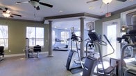 This fitness center is a thoughtfully designed single-floor space that combines functionality with an inviting architectural aesthetic. The room is anchored by a series of decorative columns that lend a classic, structured feel to the interior, complementing the soft sage-green walls and light blue accent tones throughout. A ceiling fan overhead provides comfortable air circulation during workouts, while recessed lighting ensures the space remains well-lit at all times.

The cardio equipment area is generously outfitted with multiple treadmills, elliptical machines, and a stair climber, all arranged to allow comfortable spacing between users. Large arched and rectangular windows line the walls, flooding the space with natural light and offering pleasant outdoor views of surrounding trees — creating an open, airy atmosphere that enhances the overall workout experience.

The flooring features a durable, exercise-appropriate carpet that helps define the workout zones within the space. A dedicated strength and stretching area with a bench and additional equipment rounds out the facility. The overall ambiance is clean, bright, and welcoming, making this fitness center a practical and comfortable space suited for a full range of cardio and strength training activities.
