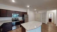 This single-floor residence offers a thoughtfully designed open-concept layout that seamlessly connects the kitchen and living areas. The kitchen is a standout feature, showcasing rich dark-wood cabinetry extending to the ceiling, light granite countertops, and a curved center island that provides additional prep space and casual seating. A full suite of stainless steel appliances — including a refrigerator, range, microwave, and dishwasher — completes the space. Durable hardwood-style flooring runs throughout the main living and kitchen areas, contributing to a clean, cohesive aesthetic.

The living area is open and airy, benefiting from a bank of windows fitted with blinds that allow for abundant natural light while maintaining privacy. The bedrooms are carpeted for comfort, each featuring windows with outdoor views and ample wall space. One bedroom includes a ceiling fan with light fixture. The bathroom is finished with matching dark-wood cabinetry, a granite-topped vanity with an undermount sink, a large framed mirror, and a full soaking tub with shower. Recessed lighting throughout the unit provides consistent, modern illumination. The space presents a clean, contemporary design with a neutral color palette and quality finishes throughout.