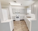 This single-story residential unit spans 720 square feet and features 3 bedrooms, 1 bathroom, a kitchen, laundry room, entrance, and hallway. The three bedrooms are finished with neutral gray carpet and soft gray walls, offering a clean, understated aesthetic. The primary bedroom measures 250 square feet and includes a built-in double-door closet. Two additional bedrooms feature windows with views of surrounding greenery, providing natural light throughout. A ceiling fan with light fixture is installed in the central living area near the entrance hallway. The kitchen is outfitted with gray shaker-style cabinetry, light stone-pattern countertops, wood-look vinyl flooring, a white electric range with overhead ventilation, a double stainless steel sink, a dishwasher, and ample upper and lower cabinet storage. The bathroom features wood-look vinyl flooring, a white vanity with gray-framed mirror, chrome towel bars, and a tub/shower combination. The dedicated laundry room includes wire shelving, washer/dryer hookups, and wood-look vinyl flooring. Neutral tones and practical finishes are consistent throughout, creating a cohesive and functional living environment.
