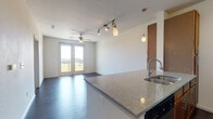 This single-floor unit offers a well-designed open-concept layout that seamlessly connects the kitchen and living areas. The kitchen is equipped with warm-toned wood cabinetry, granite countertops, a stainless steel refrigerator, range, and microwave, complemented by a peninsula island with pendant lighting overhead. Track lighting runs throughout the main living space, adding a modern touch alongside ceiling fans that promote comfortable airflow. Dark hardwood-style flooring flows continuously through the open living area, creating a cohesive and polished look. French doors in the living space invite abundant natural light and lead out to a balcony, offering open views of the surrounding landscape. The bedroom features plush carpeting, a ceiling fan, and a window fitted with blinds that allows for controlled natural light. The bathroom is finished with clean white tile and includes a glass-enclosed walk-in shower with brushed nickel fixtures, a pedestal sink, and a toilet. The overall design is clean and functional, with a neutral color palette of whites and soft grays throughout, providing a versatile backdrop suited to a variety of interior styles.