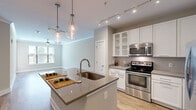 Located at 2875 Crescent Pkwy in Atlanta, GA, this one-bedroom, one-bathroom residence offers a smartly designed floor plan that maximizes every square foot. The open-concept kitchen and living area serves as the heart of the home, anchored by a large center island with a quartz countertop, an under-mount sink, and elegant glass pendant lighting overhead. The kitchen is outfitted with white cabinetry, stainless steel appliances including a refrigerator, electric range, and microwave, as well as a classic tile backsplash. Track lighting adds a modern touch throughout. Warm wood-look flooring extends across the main living area, creating a cohesive and inviting atmosphere. Natural light filters in through multiple windows, offering views of the surrounding greenery. The bedroom features plush carpeting, a ceiling fan, and generous window placement that fills the room with natural light. The bathroom connects to a dedicated walk-in closet with built-in shelving, providing practical storage. Tile flooring, white vanity cabinetry, and quartz countertops complete the bathroom. Crown molding and neutral tones throughout lend the space a clean, contemporary aesthetic ideal for comfortable everyday living.