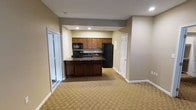 This single-floor unit is designed with an open-concept layout that seamlessly connects the living area to the kitchen, creating a spacious and functional flow throughout. The kitchen is well-appointed with rich dark wood cabinetry, dark granite-style countertops, and black appliances, including a refrigerator, range, and microwave. A peninsula-style counter serves as a natural divider between the kitchen and the main living space, providing additional prep and dining space. Recessed lighting in the kitchen area ensures ample illumination, while the neutral gray and beige wall tones contribute to a calm, cohesive atmosphere. The living area is finished with a patterned, wall-to-wall carpet in warm neutral tones, adding comfort underfoot. White trim and panel-style doors frame the space neatly, lending a classic and clean aesthetic. Multiple doorways lead to additional rooms, suggesting a thoughtful and practical floor plan. Overall, the unit offers a well-organized, move-in-ready environment suited for everyday living, with a consistent neutral palette that allows for versatile interior styling.