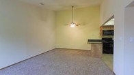 This single-story space showcases an open-concept layout that seamlessly connects the living area to the kitchen. The living area features wall-to-wall carpeting in a neutral tone, creating a comfortable and cohesive foundation throughout the space. A ceiling fan with an integrated light fixture is centrally mounted, providing both air circulation and ambient lighting while adding a functional design element to the room.

The kitchen is positioned adjacent to the living area and is defined by a raised breakfast bar with dark countertops, offering a natural division between the two spaces while maintaining an open feel. Light wood cabinetry lines the kitchen walls, complemented by black appliances, including a built-in microwave and a freestanding range, creating a classic contrast. The smooth tile flooring in the kitchen distinguishes it from the carpeted living area.

The walls throughout are painted in a soft, warm yellow-green hue, contributing to a bright and welcoming atmosphere. Overall, the space offers a functional, open design well-suited for everyday living and casual entertaining.