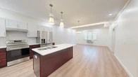 This thoughtfully designed 2-bedroom, 1-bathroom residence in Milford, CT offers a clean, modern aesthetic with light hardwood-style flooring and crisp white walls throughout. The open-concept kitchen and living area serves as the heart of the home, anchored by a striking kitchen island with a double-basin sink, rich dark mahogany cabinetry on the lower level, and bright white upper cabinets. A herringbone tile backsplash, stainless steel appliances — including a French door refrigerator and range — and elegant pendant lighting complete the well-appointed kitchen. The living area flows seamlessly from the kitchen, featuring a tray ceiling with crown molding and recessed lighting, adding a touch of architectural refinement. The primary bedroom is generously sized with a tray ceiling, crown molding, recessed lighting, and a triple window with blinds that brings in natural light. The second bedroom is a comfortable, light-filled room with a window and wall-unit climate control. The bathroom is finished with marble-look tile flooring, white cabinetry, a quartz countertop, and a sleek glass-enclosed walk-in shower with marble-style surround. A hallway and dedicated closet round out this well-organized and stylishly finished residence.