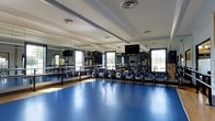 Located at 5730 Silver Falls St in Dublin, Ohio, this single-floor fitness facility is purpose-built for group exercise and cycling classes. The standout feature of the space is its expansive cycling studio, outfitted with a full fleet of stationary bikes arranged in organized rows across a vibrant blue sport flooring that is both durable and easy to maintain. Floor-to-ceiling mirrors line the front and side walls, creating a sense of openness and allowing participants to monitor their form throughout workouts. Large windows along the perimeter flood the room with natural light, contributing to an energizing and motivating atmosphere. Overhead, the ceiling features a clean, structured layout with recessed ventilation and lighting fixtures to ensure proper airflow and visibility during high-intensity sessions. Industrial-style fans are mounted strategically to provide additional cooling comfort. A wall-mounted flat-screen display is positioned for clear sightlines from all areas of the room, supporting instructor-led classes and virtual programming. The overall design of the space prioritizes functionality and performance, with thoughtful attention to the needs of fitness enthusiasts. The bright, well-ventilated environment makes this studio an inviting destination for cycling and group fitness activities in the Dublin community.