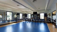 Located at 5730 Silver Falls St in Dublin, Ohio, this single-floor fitness facility is purpose-built for group exercise and cycling classes. The standout feature of the space is its expansive cycling studio, outfitted with a full fleet of stationary bikes arranged in organized rows across a vibrant blue sport flooring that is both durable and easy to maintain. Floor-to-ceiling mirrors line the front and side walls, creating a sense of openness and allowing participants to monitor their form throughout workouts. Large windows along the perimeter flood the room with natural light, contributing to an energizing and motivating atmosphere. Overhead, the ceiling features a clean, structured layout with recessed ventilation and lighting fixtures to ensure proper airflow and visibility during high-intensity sessions. Industrial-style fans are mounted strategically to provide additional cooling comfort. A wall-mounted flat-screen display is positioned for clear sightlines from all areas of the room, supporting instructor-led classes and virtual programming. The overall design of the space prioritizes functionality and performance, with thoughtful attention to the needs of fitness enthusiasts. The bright, well-ventilated environment makes this studio an inviting destination for cycling and group fitness activities in the Dublin community.