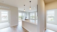 This well-appointed single-floor residence showcases a thoughtfully designed open-concept layout that seamlessly connects the kitchen, dining, and living areas. The kitchen is a standout feature, equipped with rich dark wood cabinetry, a full suite of stainless steel appliances including a refrigerator, electric range, and over-the-range microwave, and a crisp white quartz peninsula with an undermount sink and matte black faucet. Stylish pendant lighting hangs above the peninsula, adding a modern touch to the space. Light wood-tone flooring runs throughout the main living areas, contributing to a bright and airy atmosphere. The living area is flooded with natural light through large windows and a sliding glass door that opens to a private balcony, offering open outdoor views of the surrounding landscape. The bedroom features plush carpeting, oversized windows that frame expansive views, and direct access to adjacent rooms, providing a comfortable and connected feel. The bathroom is finished with a white quartz vanity countertop, an undermount sink, a bold black-framed mirror, and a sliding glass shower enclosure. Throughout the space, neutral tones, clean lines, and modern fixtures create a cohesive and contemporary aesthetic.