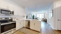 This single-floor residence showcases a thoughtfully designed open-concept layout that seamlessly connects the kitchen and living areas. The kitchen is outfitted with stainless steel appliances, including a French-door refrigerator, electric range, over-the-range microwave, and dishwasher. Light wood cabinetry and white upper cabinets pair with white quartz countertops, while a peninsula with a sink and dual pendant lights above serves as a natural dividing point between the kitchen and living space. Wood-look flooring runs throughout the main living areas, lending warmth and continuity. The living room benefits from a large window that invites generous natural light and urban views, complemented by a ceiling fan overhead. Both bedrooms feature plush carpeting, large windows with horizontal blinds and curtain panels, and ample space for furnishings. The bathroom is finished with wood-look flooring, a single-sink vanity with light cabinetry and quartz countertop, open shelving, and a tub-shower combination with a marble-style surround. Recessed lighting and a clean, neutral color palette of whites and soft naturals give the entire space a contemporary, cohesive aesthetic.