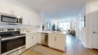 This single-floor residence showcases a thoughtfully designed open-concept layout that seamlessly connects the kitchen and living areas. The kitchen is outfitted with stainless steel appliances, including a French-door refrigerator, electric range, over-the-range microwave, and dishwasher. Light wood cabinetry and white upper cabinets pair with white quartz countertops, while a peninsula with a sink and dual pendant lights above serves as a natural dividing point between the kitchen and living space. Wood-look flooring runs throughout the main living areas, lending warmth and continuity. The living room benefits from a large window that invites generous natural light and urban views, complemented by a ceiling fan overhead. Both bedrooms feature plush carpeting, large windows with horizontal blinds and curtain panels, and ample space for furnishings. The bathroom is finished with wood-look flooring, a single-sink vanity with light cabinetry and quartz countertop, open shelving, and a tub-shower combination with a marble-style surround. Recessed lighting and a clean, neutral color palette of whites and soft naturals give the entire space a contemporary, cohesive aesthetic.