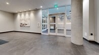 Located at 169 Rutland North Road in Kelowna, BC, this single-floor hallway space offers a thoughtfully designed common area that balances functionality with contemporary style. The layout follows an L-shaped configuration, providing distinct zones that serve different practical purposes while maintaining a cohesive visual identity throughout.

The flooring is finished in large-format grey stone-look tiles, lending the space a clean, modern character that carries consistently from the entrance through to the elevator corridor. Concrete-finish cylindrical columns punctuate the main lobby area, adding an industrial-chic architectural element that complements the neutral palette of soft white and warm grey walls.

The lobby area is furnished with wooden benches and a statement tropical plant in a white vessel, positioned in front of a mirror-and-dark-slat accent wall. Framed artwork adds a curated touch. Recessed pot lighting throughout ensures the space is well-lit without feeling clinical.

A smart parcel locker system is integrated directly into the hallway, offering a practical, modern solution for package management. The elevator bank features two stainless steel elevator doors accompanied by a runner mat, with a secondary fire exit door visible beyond. Glass entry doors connect to an exterior vestibule, allowing natural light to filter into the entrance zone.