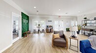 Located at 7959 Cottage Hill Rd in Mobile, Alabama, this single-floor space offers a cohesive open-concept layout combining a fully equipped kitchen and a spacious living area. Light oak vinyl plank flooring flows throughout, unifying the space and contributing to its bright, airy atmosphere.

The kitchen is outfitted with granite countertops, white cabinetry, a stainless steel refrigerator, and a peninsula with bar seating — ideal for casual dining or entertaining. Multiple windows fitted with white shutters invite natural light while maintaining privacy.

The living area is generously proportioned and features a tray ceiling with recessed lighting that adds depth and warmth to the room. Crown molding runs throughout, lending an elegant architectural touch. French doors along one wall provide an outdoor view and flood the interior with additional natural light. Wall space is utilized artfully, with framed abstract artwork and decorative wall panels enhancing the contemporary aesthetic.

A standout feature is a striking living green wall display branded with the property's identity, complemented by a dedicated coffee bar area, suggesting a welcoming, community-oriented atmosphere. The overall ambiance is clean, modern, and well-appointed, making this space both functional and visually engaging.