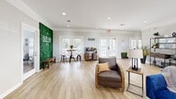 Located at 7959 Cottage Hill Rd in Mobile, Alabama, this single-floor space offers a cohesive open-concept layout combining a fully equipped kitchen and a spacious living area. Light oak vinyl plank flooring flows throughout, unifying the space and contributing to its bright, airy atmosphere.

The kitchen is outfitted with granite countertops, white cabinetry, a stainless steel refrigerator, and a peninsula with bar seating — ideal for casual dining or entertaining. Multiple windows fitted with white shutters invite natural light while maintaining privacy.

The living area is generously proportioned and features a tray ceiling with recessed lighting that adds depth and warmth to the room. Crown molding runs throughout, lending an elegant architectural touch. French doors along one wall provide an outdoor view and flood the interior with additional natural light. Wall space is utilized artfully, with framed abstract artwork and decorative wall panels enhancing the contemporary aesthetic.

A standout feature is a striking living green wall display branded with the property's identity, complemented by a dedicated coffee bar area, suggesting a welcoming, community-oriented atmosphere. The overall ambiance is clean, modern, and well-appointed, making this space both functional and visually engaging.