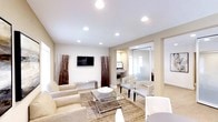 This thoughtfully designed single-floor space exudes a clean, modern aesthetic anchored by a warm neutral color palette throughout. The main lounge area serves as the centerpiece, furnished with plush beige seating arranged around a sleek glass-and-chrome coffee table atop a richly textured area rug. Recessed ceiling lighting provides consistent, even illumination across the space, enhancing its bright and polished atmosphere. A wall-mounted flat-screen television, flanked by tall decorative dried grass arrangements, adds both function and visual interest to the lounge. Adjacent to the seating area, a built-in workstation offers a dedicated space for computer use, keeping productivity seamlessly integrated into the environment. A pair of elegantly patterned accent chairs provides additional seating nearby, creating a comfortable conversational nook. Toward the rear, a private office or meeting room is enclosed with frosted glass partitioning, allowing natural light to filter through while maintaining privacy. The use of glass and clean architectural lines throughout the space gives it a professional yet inviting character, making it well-suited for a professional office, salon reception, or client-facing business environment.