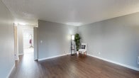 This single-floor space presents a thoughtfully designed interior that balances simplicity with comfort. The living area is defined by rich, dark hardwood flooring that runs throughout, providing a warm and grounding contrast to the cool, neutral gray walls. The textured ceiling adds subtle visual interest while maintaining a clean, understated aesthetic overall.

The open layout of the main living area offers generous room to move and arrange furnishings to suit a variety of purposes. Crisp white baseboards frame the perimeter of the room, adding a polished finish to the space. A hallway visible from the main area suggests additional rooms beyond, providing a sense of depth and connectivity throughout the floor plan.

Bright and airy in feel, the space benefits from well-placed lighting that enhances its inviting atmosphere. The neutral color palette throughout creates a versatile backdrop that adapts easily to a range of interior styles and décor preferences.

Overall, this single-level space offers a clean, functional, and welcoming environment. Its combination of durable hardwood flooring, soft gray tones, and an open, uncluttered layout makes it a practical and appealing setting suitable for everyday living.