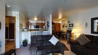 This single-floor residence is designed around an open-concept floor plan that seamlessly integrates the living, dining, and kitchen areas. The living room serves as the central hub, furnished with a large, dark upholstered sectional sofa accented by a white throw pillow, and anchored by a glass-top coffee table. Neutral-toned carpeting adds warmth underfoot throughout this area. A structural column subtly defines the transition between spaces without disrupting the open flow.

The kitchen sits adjacent to the living area, separated by a raised breakfast bar with barstool seating. It features dark wood cabinetry, a built-in microwave, and a light-colored countertop that provides ample workspace. The dining area is positioned nearby, equipped with a dark wood table and matching chairs, ideal for everyday meals.

A ceiling fan with a light fixture illuminates the central area, complementing the overhead lighting throughout. A bookcase filled with media and a floor lamp add functional character near the dining space. Decorative wall accents and a display cabinet contribute personality to the home's overall welcoming and lived-in ambiance.