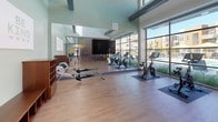 Located at 2205 Natomas Park Dr in Sacramento's Natomas Park neighborhood, this contemporary fitness center is thoughtfully designed to inspire an active lifestyle. The space is defined by soaring ceilings with exposed beams and an abundance of natural light streaming through floor-to-ceiling glass walls that frame views of the outdoor pool and surrounding community. The main cardio area features a row of treadmills positioned directly in front of the expansive windows, allowing users to enjoy the outdoor scenery during their workouts. A dedicated cycling and rowing zone includes spin bikes and rowing machines arranged on durable hardwood-style flooring, complemented by a large flat-screen display. The strength training area is finished with resilient rubber flooring and houses a cable machine, adjustable benches, and a full dumbbell rack, all set against a wall of mirrors that amplify the sense of space. A bold geometric mural in shades of navy, teal, yellow, and gray energizes one wall, adding a vibrant, motivating aesthetic. Additional amenities include a recumbent bike station, storage cubbies, and convenient access to sanitizing supplies. The seamless flow between zones and the well-curated equipment selection make this fitness center a comprehensive and welcoming facility for residents.
