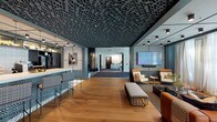 This single-floor clubhouse is a thoughtfully designed community space that balances sophisticated aesthetics with functional amenities. The entrance lobby makes a strong first impression with its slatted wood ceiling, brick fireplace, built-in bench seating, and floor-to-ceiling glass doors that invite abundant natural light and outdoor views. Warm carpeting and layered area rugs create an inviting atmosphere, while curated décor—including a guitar, sculptural animal figures, and framed artwork—adds personality and charm.

Transitioning into the main gathering area, the space features rich hardwood flooring beneath a striking geometric lattice ceiling finished in dark tones, lending dramatic contrast to the open layout. A bar-style kitchen island with patterned paneling and counter seating serves as a central hub, complemented by nearby access to a vending and refreshment area. A large-scale lounge anchors the entertainment zone, furnished with a tufted leather sofa, accent chairs, and an upholstered bench, all arranged around a wall-mounted flat-screen display. Sheer floor-length drapery softens the natural light streaming through tall windows. Additional spaces include a darkly finished room with curved architectural walls and sleek black double doors, offering a more intimate or private-use environment. Throughout, the clubhouse maintains a cohesive modern-industrial design language with high-end finishes and curated details.