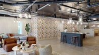 This thoughtfully designed single-floor commercial space offers a generous, open layout that balances industrial aesthetics with refined, curated interiors. Exposed ductwork, black-painted ceilings, and structural concrete columns define the raw architectural framework, while warm lighting from an array of glass pendant fixtures softens the atmosphere throughout.

The central gathering area features a large, fluted blue bar island with a dark stone countertop and wooden bar stools, serving as an anchor for the open-plan lounge. Bold, graphic botanical wallpaper creates a vibrant focal point, adding color and energy to the space. The lounge area is furnished with plush rust-toned armchairs, layered rugs, and woven accent pieces, encouraging relaxed social interaction.

A mezzanine-style overlook with wrought iron railings brings in abundant natural light through floor-to-ceiling windows, offering views of the surrounding streetscape. The hallways lead to private rooms, including a quiet meeting room with a round wood table, upholstered chairs, neutral textured walls, and framed artwork. A standout feature is a dedicated room lined with a floor-to-ceiling wood wine storage wall, displaying labeled wine crate panels under track lighting. Light wood-look tile flooring flows consistently throughout, unifying the entire space.