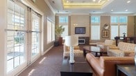 This single-floor communal space is thoughtfully designed to offer both relaxation and social functionality. The main lounge area serves as the centerpiece, featuring a coffered ceiling with warm ambient lighting, patterned carpeting, and a wall-mounted flat-screen TV flanked by a stone-accented fireplace. Generous windows and French doors flood the room with natural light while offering views to the exterior. Comfortable seating — including a leather sofa, upholstered armchairs, and a glass-top oval coffee table — is arranged to encourage conversation and leisure. An adjacent dining nook provides bistro-style high-top tables in rich dark wood paired with sleek black stools, accented by decorative artwork and tall windows that bring in additional daylight. The kitchen area is outfitted with dark espresso cabinetry, granite countertops, stainless steel appliances including a side-by-side refrigerator and microwave, under-cabinet lighting, and a large central island with bar-height seating. Dark tile flooring throughout the kitchen complements the warm tones of the lounge. The overall design palette — warm neutrals, earthy greens, and deep wood tones — creates a cohesive, welcoming atmosphere suitable for gatherings or quiet individual use.