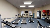 The Kennedy fitness center is a well-equipped, single-floor facility designed to support a full range of workout routines. The space is bright and functional, illuminated by recessed fluorescent panel lighting set against a textured ceiling, creating a clean and energizing atmosphere throughout.

The cardio zone features multiple treadmills, elliptical machines, and upright stationary bikes — all from True and similar brands — arranged in a row facing arched windows that allow natural light to filter in. A wall-mounted flat-screen television is positioned for easy viewing during cardio sessions.

The strength training area is equally comprehensive, housing a variety of Paramount cable and resistance machines, a multi-function cable column, lat pulldown stations, a leg press, and adjustable benches. A dedicated free weights section includes a dumbbell rack with a range of weights, along with a stability ball rack stocked with multiple ball sizes for functional training.

The flooring throughout is a durable, speckled rubberized surface well-suited for high-traffic fitness use. A bold blue accent wall energizes the strength training zone, while neutral tones dominate the cardio area. The facility includes clearly marked exits, a wall-mounted clock, and a small sink area, ensuring both safety and convenience for users.