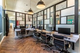 This thoughtfully designed single-floor workspace blends contemporary aesthetics with functional office planning. The most striking architectural feature is the floor-to-ceiling black steel-framed glass partition wall, which divides the space while maintaining visual openness. Frosted and clear glass panels create a sense of depth and allow natural light to filter throughout the interior.

The workstation area is anchored by a long granite-topped desk running along the partition, accommodating multiple computer stations with ergonomic black office chairs. Oversized black industrial pendant lights hang from the elevated ceiling, providing focused task lighting and contributing to the space's modern-industrial character.

Rich herringbone-patterned hardwood floors extend throughout, adding warmth and visual texture to the otherwise sleek interior. Walls are painted in a deep teal-green tone, offering a bold contrast to the white upper walls and ceilings.

Beyond the partition, an open lounge and kitchen area features an island with a dark wood base and white marble countertop, surrounded by an eclectic mix of seating in navy, red, and yellow tones. Large windows bring in natural light and outdoor views. Decorative indoor plants, wall-mounted acoustic panels, and colorful artwork complete the polished, professional ambiance.
