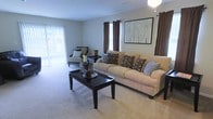 This single-floor residence centers around a spacious living room designed with a clean, neutral aesthetic. The room is finished with wall-to-wall carpeting in a warm beige tone, contributing to a comfortable and cohesive atmosphere. A large sliding glass door floods the space with natural light and provides direct access to an outdoor area, while an additional window with dark brown curtain panels adds a layered, decorative touch to the room's design. The living area accommodates generous seating, including a substantial sofa accented with patterned throw pillows in coordinating earth tones, complemented by a side chair and a bold dark-wood coffee table that serves as a focal point. Framed wall art above the seating area adds a finishing decorative element. The ceiling features a light fixture that provides ambient lighting throughout the room. The overall ambiance of the space is relaxed and well-organized, with a neutral color palette of tans, browns, and soft blues creating a welcoming, functional environment suited for everyday living and entertaining.