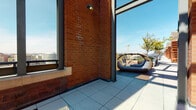 This rooftop terrace is a well-designed outdoor space that combines architectural character with functional amenities, offering an elevated urban retreat. The terrace is defined by prominent exposed brick structures featuring decorative tile accents and a curved architectural element, adding visual interest and a sense of permanence to the space.

Access to the terrace is provided through double glass doors framed in sleek metal, opening onto large-format tiled flooring that extends throughout the space. A pergola structure overhead provides partial shade to one seating area, where a cushioned sofa and woven chairs surround a round bistro table — ideal for socializing or relaxing outdoors.

A distinctive oval-shaped lounge daybed is positioned to take full advantage of the open-air setting. Potted topiaries and planters with seasonal greenery are thoughtfully distributed around the terrace, softening the hardscape and adding natural texture.

Metal railing borders the perimeter, ensuring safety while preserving unobstructed panoramic views of the surrounding cityscape, including neighboring buildings, treetops, and open sky. The terrace receives abundant natural light throughout the day, making it a bright and inviting outdoor space well-suited for both leisure and gathering.