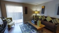 This single-floor living space is designed with a contemporary aesthetic that balances comfort and style. The living area serves as the central gathering space, showcasing rich hardwood flooring that adds warmth and visual depth throughout. Neutral, earthy-toned walls provide a versatile backdrop that complements the carefully selected furnishings.

A large sliding glass door with vertical blinds allows an abundance of natural light to filter into the room, while also providing direct access to an outdoor area. The living room is anchored by a substantial dark brown sofa, generously accented with green and patterned throw pillows that introduce a pop of color. A wooden coffee table sits atop a bold geometric-patterned area rug, adding a modern focal point to the space.

Ambient lighting is provided by a warm table lamp positioned on a console table along the back wall, which also features small decorative plants, contributing to an inviting atmosphere. Tasteful wall art in complementary tones adds a curated, intentional quality to the décor. Overall, the space feels cohesive, functional, and thoughtfully designed.