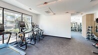 This contemporary fitness center is thoughtfully designed to support a full range of workout needs within a bright, open environment. The space is divided into distinct functional zones, each dedicated to a different category of exercise. The cardio area houses a row of treadmills and elliptical machines positioned alongside large floor-to-ceiling windows, offering expansive views of lush greenery and the surrounding streetscape — an inviting backdrop for any workout. Linear pendant lighting runs along the white ceilings, contributing to the clean, modern aesthetic throughout.

The strength training zone is comprehensively outfitted with a Matrix functional trainer, a multi-use cable machine system, and a full rack of dumbbells alongside medicine balls and various free weights. Large wall-mounted mirrors extend the visual depth of the space and are complemented by elegant vertical wood slat accents that add warmth to the otherwise crisp, minimalist interior. The flooring throughout consists of durable, dark rubberized gym flooring, well-suited for high-traffic fitness use. Recessed ventilation and modern track lighting ensure comfort and visibility across the entire facility. The space is accessible via a keyed entry door and includes clearly marked emergency exits, reflecting a practical and safety-conscious design.