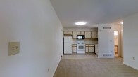 This single-floor residence offers a practical and open layout that seamlessly connects the living area to the kitchen, creating a unified and functional space. The living area features neutral-toned carpeting that adds warmth underfoot, while the clean white walls provide a bright, airy atmosphere throughout.

The kitchen is well-equipped and designed for everyday use, featuring cream-colored cabinetry that extends to the upper walls for ample storage. Appliances include a full-size white refrigerator, a white electric range with oven, a built-in microwave above the range, and a dishwasher. The kitchen flooring transitions to a smooth, light-toned surface, distinguishing it from the carpeted living area.

Overhead flush-mount lighting illuminates the kitchen, while the open sightlines between the two spaces ensure a cohesive and inviting environment. Visible HVAC vents integrated into the walls and floor indicate a central heating and cooling system throughout the unit. White baseboards and clean wall finishes contribute to a well-maintained, move-in-ready aesthetic. Overall, the space is designed with simplicity and practicality in mind, making it a comfortable and efficient single-floor living environment.
