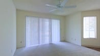 This single-floor space offers a clean, functional layout designed with comfort in mind. The main living area is finished with wall-to-wall carpeting in a neutral tone, creating a soft and welcoming underfoot feel that complements the room's light color palette. Pale yellow walls reflect natural light throughout, contributing to a bright and airy atmosphere.

A prominent feature of the space is the expansive sliding glass door spanning much of one wall, dressed with full-length vertical blinds that provide both privacy and light control. When open, this door floods the room with natural light and offers access to the outdoors. A ceiling fan is mounted overhead, offering year-round comfort and efficient air circulation.

The textured ceiling adds subtle architectural character, while multiple electrical outlets are thoughtfully positioned along the walls for practical use. The overall design is straightforward and versatile, providing a blank canvas that can accommodate a variety of furnishing arrangements and personal styles. The combination of natural light, neutral finishes, and functional fixtures makes this a comfortable and adaptable living space.