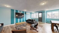 The party room at 5501 Avenue Adalbert in Côte-Saint-Luc is a well-appointed communal space designed to accommodate a variety of social gatherings. The single-floor layout is divided into distinct functional zones, offering both a relaxed lounge area and a more structured dining section.

The lounge zone features warm hardwood-style flooring, bold teal and light blue striped accent walls, and a wall-mounted flat-screen TV, creating a lively yet comfortable atmosphere. Leather seating, including a sofa and accent chairs, is arranged around a rustic wood coffee table. A wall-mounted split air conditioning unit ensures year-round comfort, complemented by baseboard heating along the perimeter.

The dining area transitions to tile flooring and is bathed in natural light from large sliding windows overlooking the surrounding neighbourhood. Bold teal walls add a vibrant contrast, and sturdy wood dining tables with mixed industrial-style seating accommodate multiple guests comfortably.

A compact kitchenette, distinguished by a hexagonal-tile clad column, stainless steel refrigerator, sink, and bar-height counter, provides practical amenities for entertaining. A textured ceiling with recessed lighting ties the space together. The room is accessed through a dedicated entrance, clearly marked as the party room.