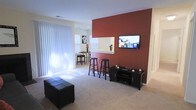 This single-story space is designed with an open-concept layout that promotes a comfortable and functional living environment. The living area is anchored by a striking deep red accent wall, which serves as a focal point and adds a warm, bold contrast to the surrounding neutral white walls and light beige carpeting. A wall-mounted flat-screen television is positioned on the accent wall, complemented by a compact media console below for storage.

The living space features large vertical blinds covering what appears to be a sliding glass door or oversized window, allowing for ample natural light to filter in while maintaining privacy. A tufted black ottoman adds a practical seating element to the room.

Transitioning toward the back of the space, a white breakfast bar with a pass-through opening connects the living area to the kitchen beyond, creating a natural division while maintaining visual openness. Two black bar stools are positioned at the counter, offering casual seating. Adjacent to the bar, a small dining area accommodates a bistro-style table and chairs, benefiting from the natural light provided by a nearby window. The overall ambiance is modern and clean.