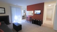 This single-story space is designed with an open-concept layout that promotes a comfortable and functional living environment. The living area is anchored by a striking deep red accent wall, which serves as a focal point and adds a warm, bold contrast to the surrounding neutral white walls and light beige carpeting. A wall-mounted flat-screen television is positioned on the accent wall, complemented by a compact media console below for storage.

The living space features large vertical blinds covering what appears to be a sliding glass door or oversized window, allowing for ample natural light to filter in while maintaining privacy. A tufted black ottoman adds a practical seating element to the room.

Transitioning toward the back of the space, a white breakfast bar with a pass-through opening connects the living area to the kitchen beyond, creating a natural division while maintaining visual openness. Two black bar stools are positioned at the counter, offering casual seating. Adjacent to the bar, a small dining area accommodates a bistro-style table and chairs, benefiting from the natural light provided by a nearby window. The overall ambiance is modern and clean.