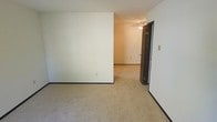 This single-floor residential space offers a clean, straightforward layout designed for comfortable everyday living. The interior is finished with wall-to-wall carpeting in a warm beige tone throughout, providing a soft and cohesive feel underfoot across the rooms. Neutral white walls keep the space feeling bright and open, offering a versatile backdrop that adapts well to a variety of furnishing styles and décor choices.

The rooms are connected through a practical corridor layout, with a doorway visible between the main living area and an adjoining room, allowing for a natural flow through the space. Dark baseboards provide a subtle contrast against the light-toned walls and carpet, adding a grounding visual element to the interior.

A ceiling fan with a light fixture is installed in the adjoining room, contributing to both air circulation and ambient lighting. A light switch is conveniently positioned near the entryway between rooms. The overall ambiance is understated and functional, making this a practical space well-suited for a range of uses. The clean lines, neutral palette, and consistent flooring throughout contribute to a unified and easy-to-maintain interior environment.
