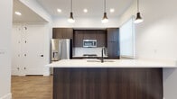 This contemporary single-floor residence showcases a thoughtfully designed open-concept layout, where clean lines and a cohesive modern aesthetic tie each space together. The kitchen serves as the centerpiece, featuring rich dark wood cabinetry that extends floor to ceiling, providing generous storage alongside a striking visual contrast against the crisp white countertops and bright walls. A spacious island anchors the kitchen, equipped with a built-in sink fitted with a matte black faucet and illuminated by a trio of stylish pendant lights overhead. Stainless steel appliances — including a French door refrigerator, gas range, and over-the-range microwave — complete the kitchen's polished, functional appeal. A geometric white tile backsplash adds a subtle textural detail. The open living area flows naturally from the kitchen, bathed in natural light from oversized windows dressed with roller shades. Warm hardwood-style flooring runs throughout the main living spaces, grounding the bright, white-painted interior. The bedroom offers plush carpeting, a walk-in closet with built-in shelving, and a window overlooking the exterior. The bathroom is finished with the same dark floating vanity, a white countertop, matte black fixtures, a large mirror, and coordinating wood-look flooring — maintaining a consistent, contemporary style throughout.